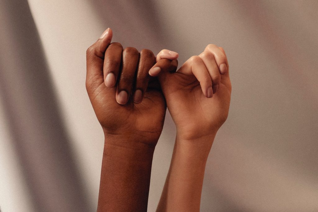 The image shows two hands interlocking their pinky fingers. One hand has a darker skin tone, while the other has a lighter skin tone. The fingers are gently held together against a soft, blurred background, conveying a sense of connection and unity.