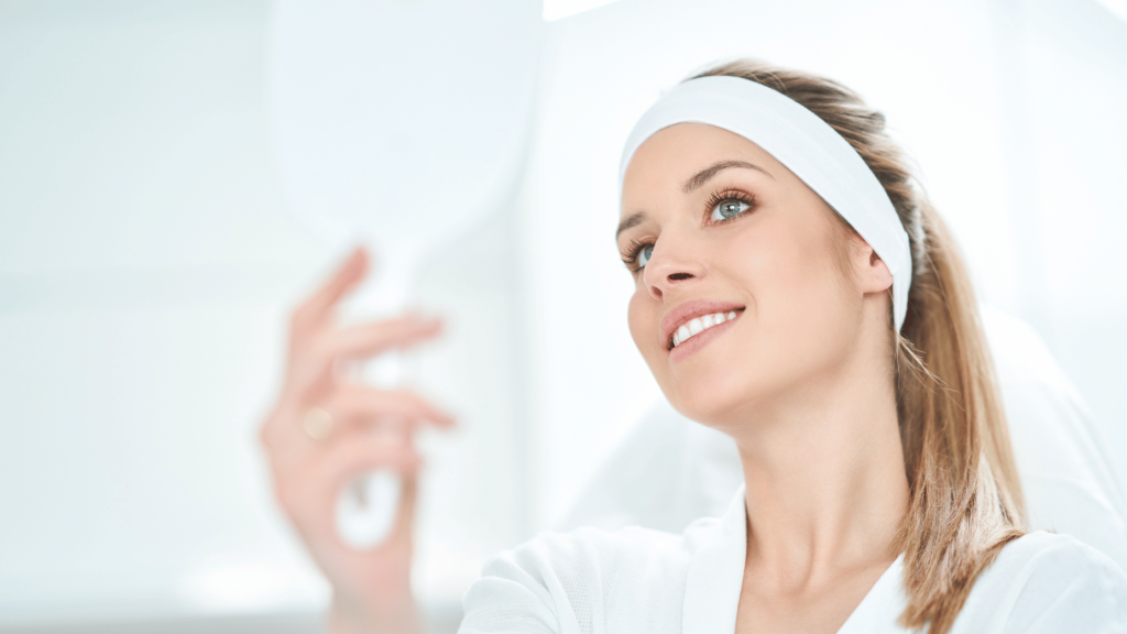 A woman standing in front of a mirror, gently touching her face while examining her reflection.